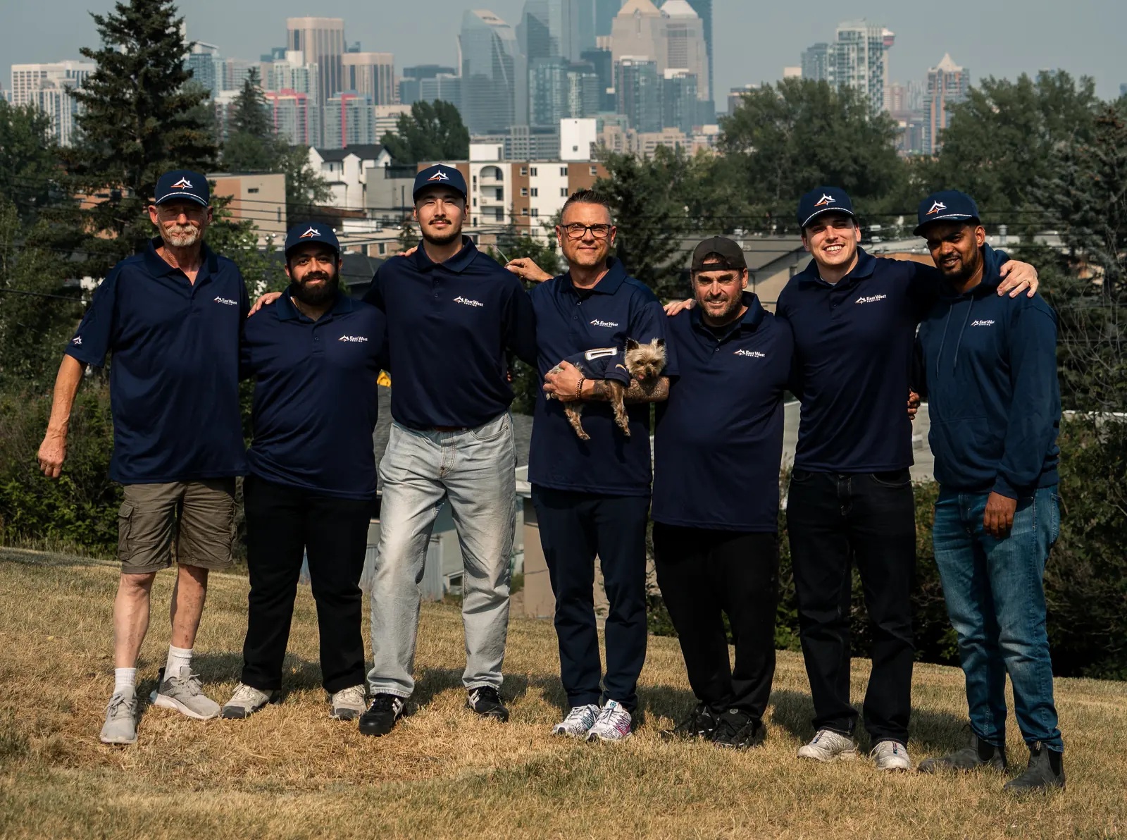 The East West Roofing team in branded uniform on a hillside with the downtown Calgary skyline behind them